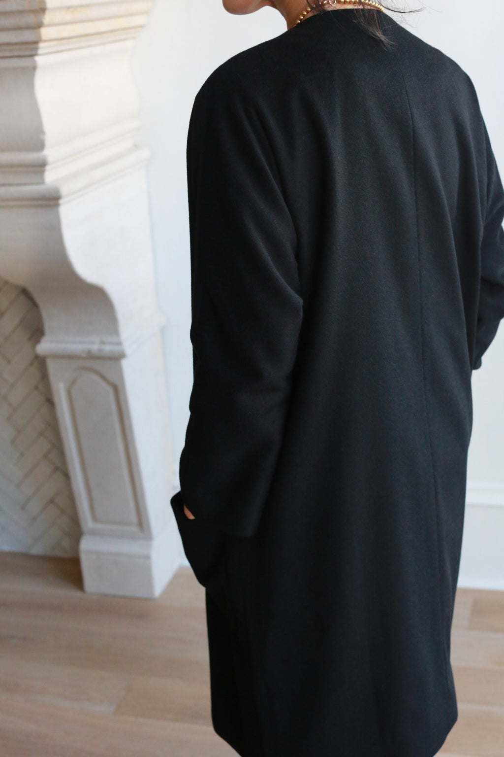 Wearing the Battersea, a long black cashmere coat with raglan sleeves, a person stands indoors by a white fireplace on light wood floors—their back mostly to the camera and only part of their head and body visible.