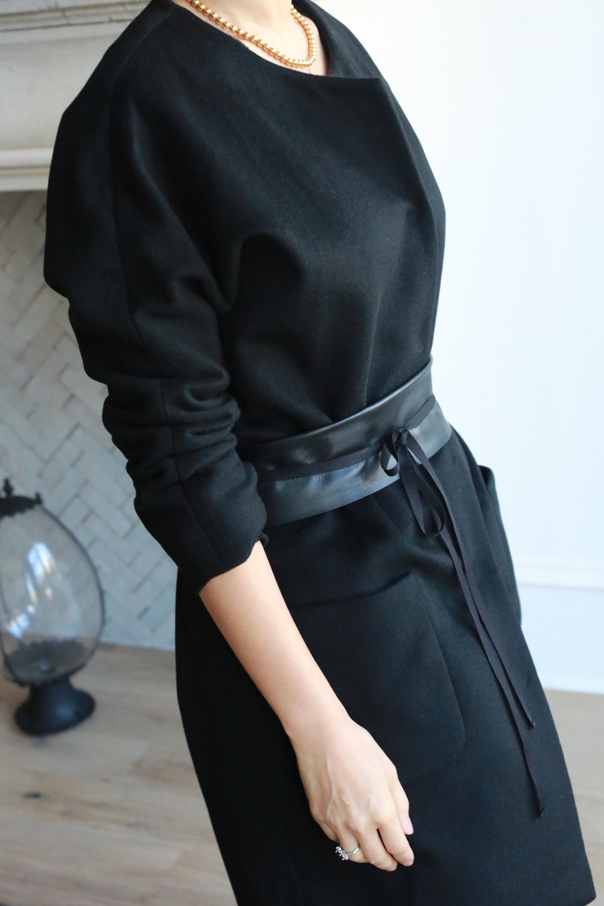 Someone wearing the Battersea black long-sleeve dress with handmade snap buttons and a black leather waist belt, accessorized with a gold necklace, stands indoors by a light herringbone-patterned wall.