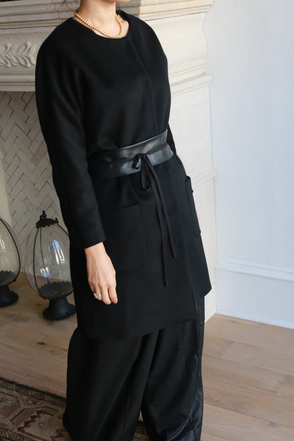 A person wearing the Battersea black cashmere coat with raglan sleeves, black wide-leg pants, and a gold necklace stands indoors near a light-colored fireplace. Their face is partially out of frame.