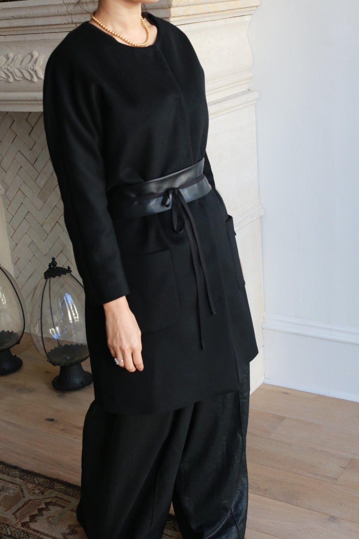 A person wearing the Battersea black cashmere coat with raglan sleeves, black wide-leg pants, and a gold necklace stands indoors near a light-colored fireplace. Their face is partially out of frame.