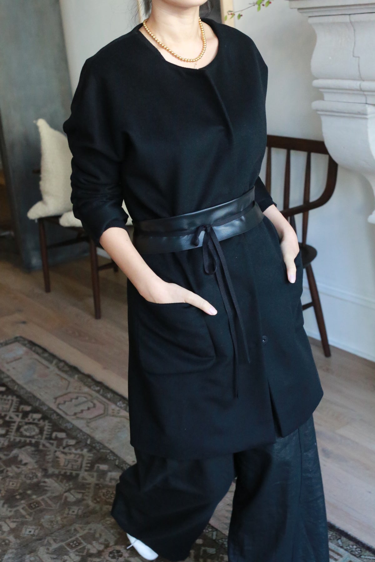 A person stands indoors with hands in the pockets of a long black Battersea cashmere coat featuring handmade snap buttons, paired with black wide-leg pants and a gold chain necklace. The face is partially out of frame; furniture and a patterned rug are visible.