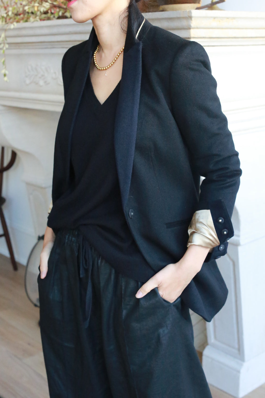 A person dressed in a stylish, casual black ensemble stands with hands in pockets. They wear the Sloane blazer with structured shoulders and rolled-up sleeves revealing a gold inner lining, a black V-neck top, and loose-fitting black pants. A gold chain necklace completes the look.