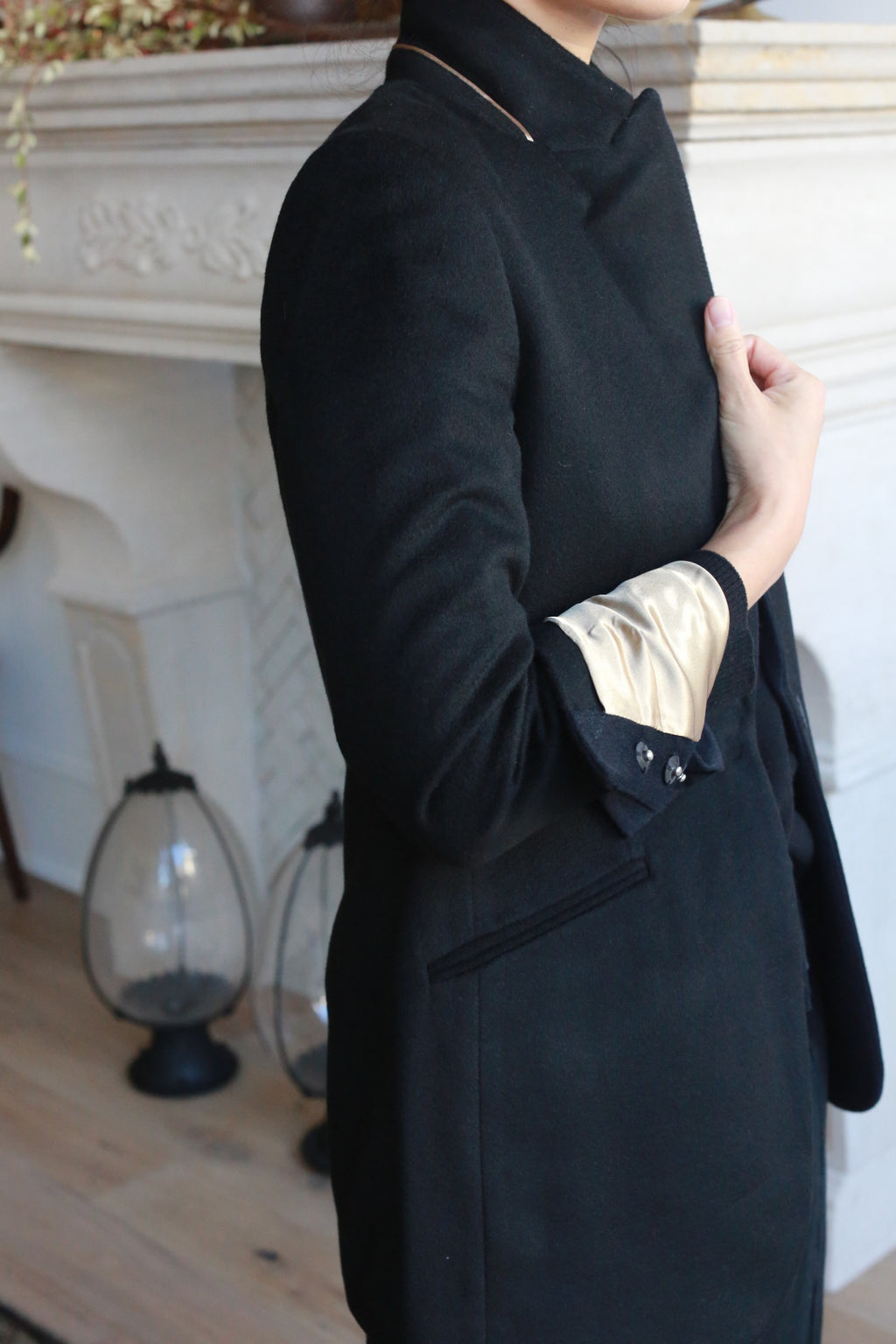 A person wearing the Sloane coat, which features an equestrian length and light-colored inner cuffs, stands indoors, partially facing the camera. In the background, there is a decorative fireplace and two lanterns on the wooden floor. The setting appears elegant and stylish.