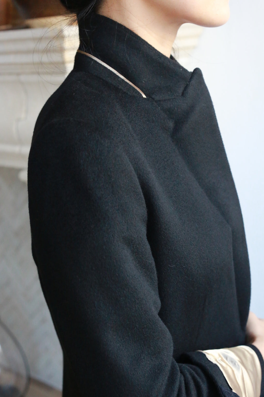 A person wearing a Sloane coat with structured shoulders, facing away from the camera, and their head turned slightly to the side. The background features a white textured wall and part of a mantle. Soft lighting highlights the woven cashmere texture of the coat.