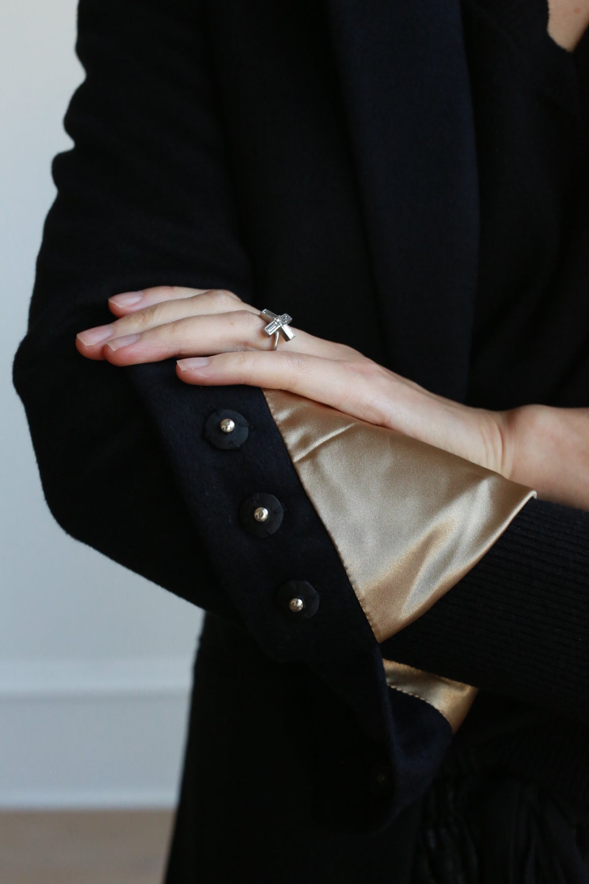 A person in a black Sloane equestrian-length coat with golden silk lining holds their hand against their chest, revealing a silver ring with a large, clear gemstone on their finger. The woven cashmere Sloane coat features three black buttons on the cuff and structured shoulders.