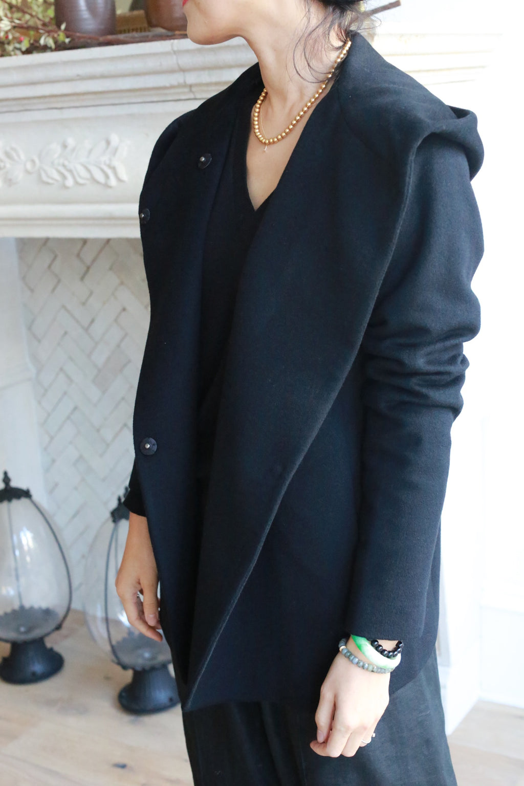 Wearing the Mitchley jacket and a gold beaded necklace, a person stands indoors by a white fireplace with decorative tiles, their face partially out of view.