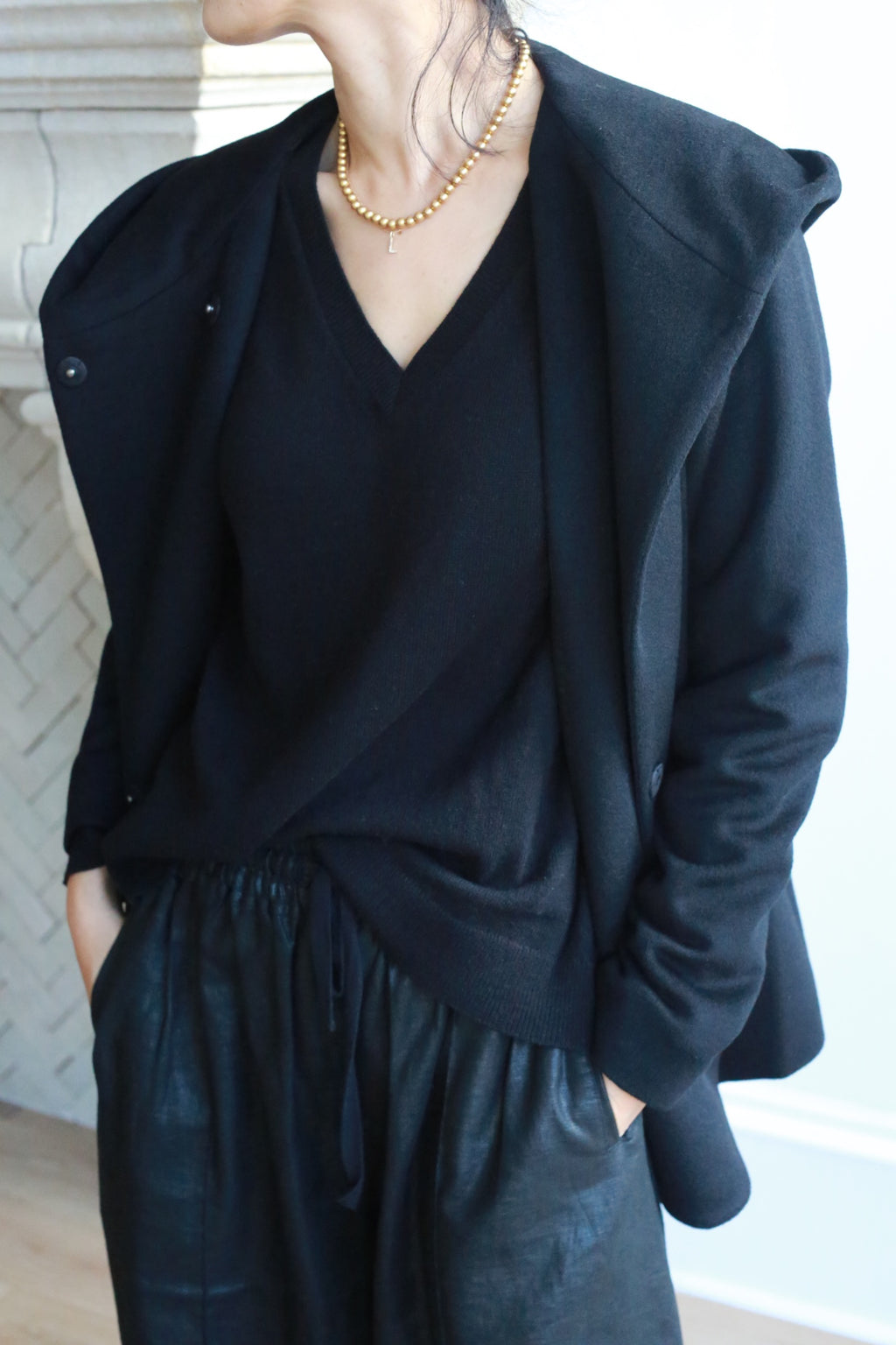 Wearing the Mitchley black V-neck sweater, an elegant jacket draped over their shoulders, black drawstring pants, and a gold beaded necklace, the person stands with hands in pockets, face partially out of frame.