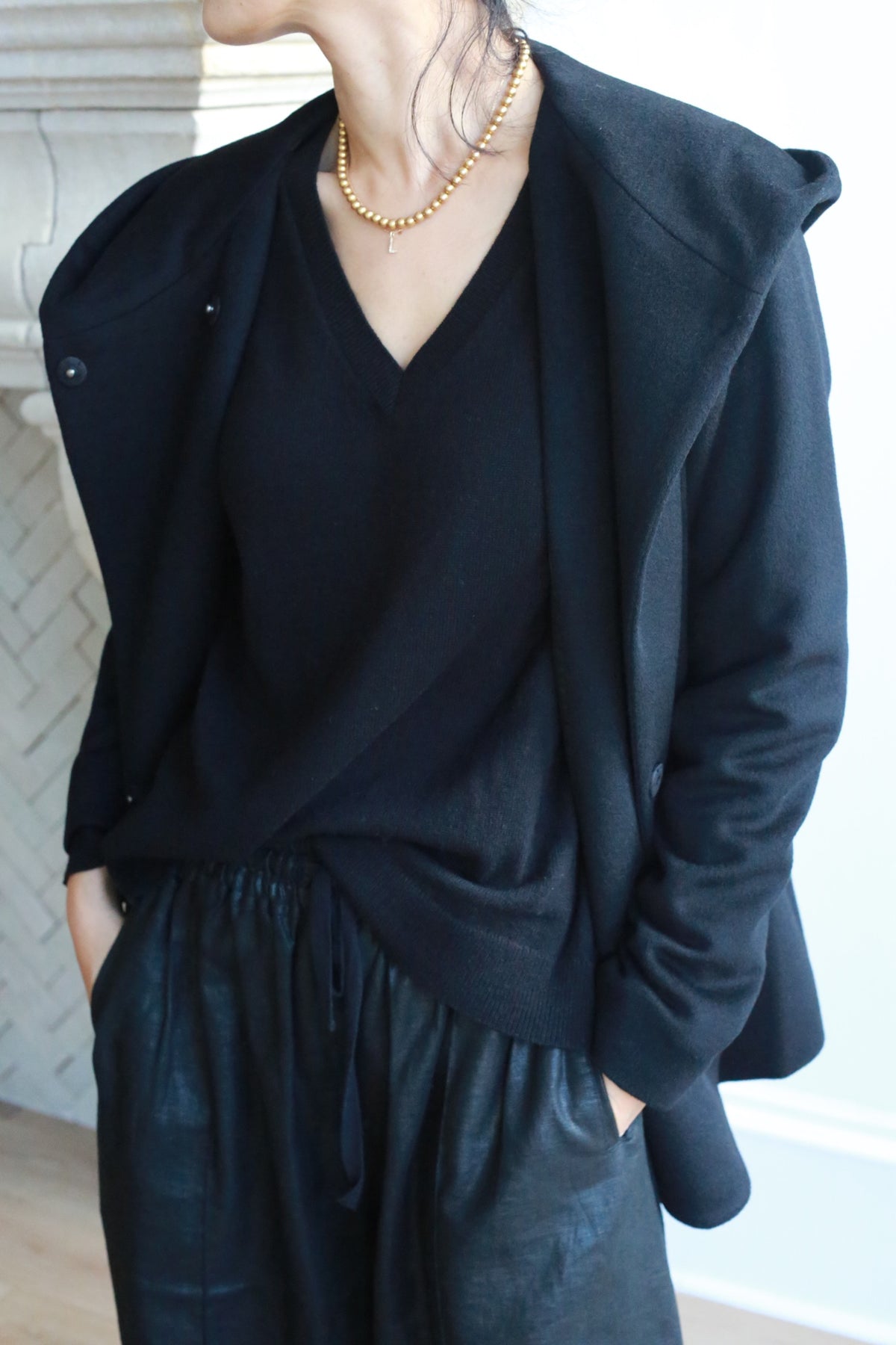 Wearing the Mitchley black V-neck sweater, an elegant jacket draped over their shoulders, black drawstring pants, and a gold beaded necklace, the person stands with hands in pockets, face partially out of frame.
