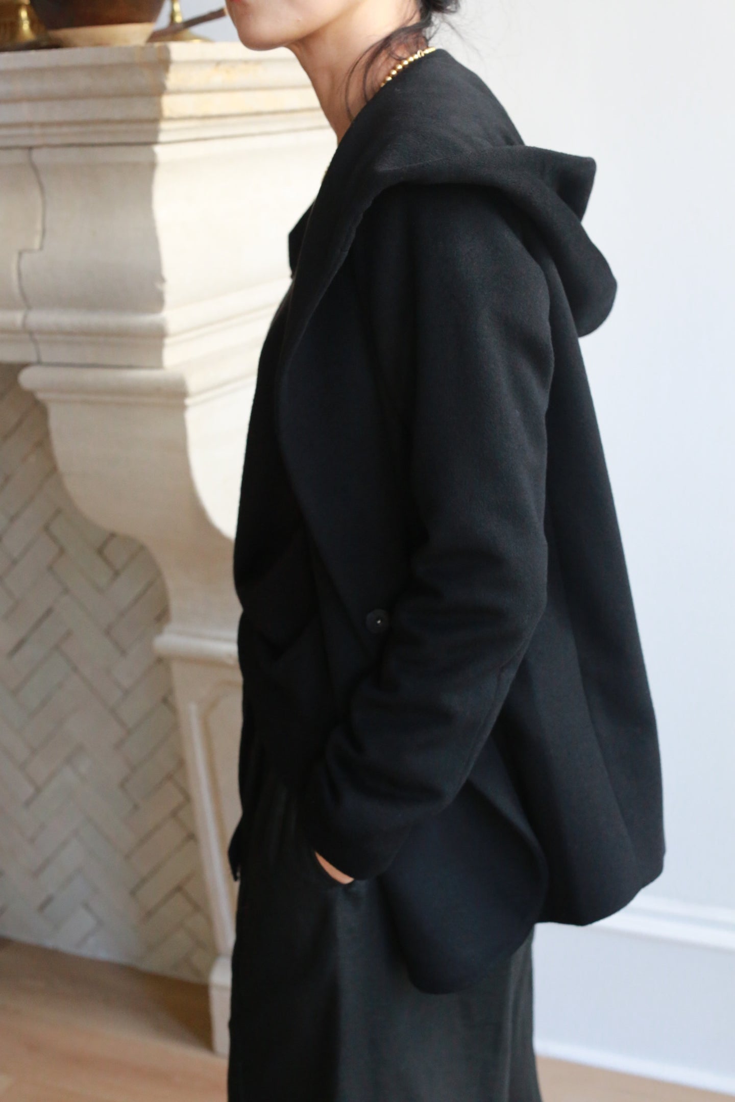 Wearing the Mitchley jacket with a cashmere hood and black pants, a person stands sideways by an ornate fireplace, one hand in their pocket and their face partially out of view.