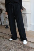 Person standing indoors in front of a white fireplace, wearing Cashmere Trousers with a drawstring and white shoes. Part of an ornate rug is visible on the wooden floor. Upper body is mostly cropped out of the frame.