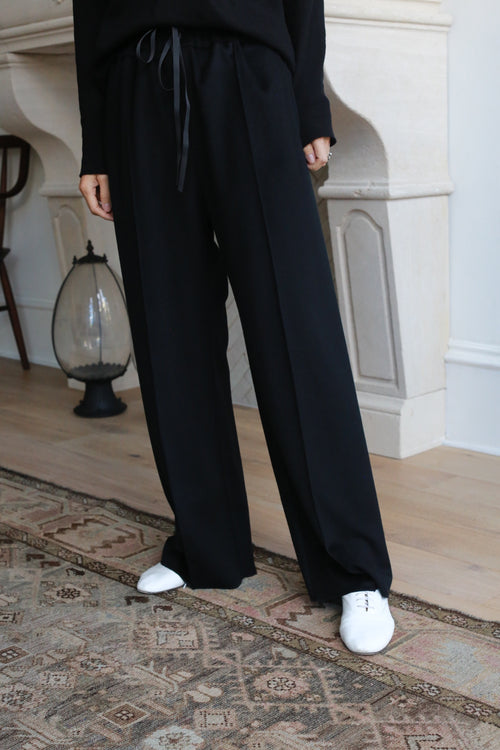 Person standing indoors in front of a white fireplace, wearing Cashmere Trousers with a drawstring and white shoes. Part of an ornate rug is visible on the wooden floor. Upper body is mostly cropped out of the frame.