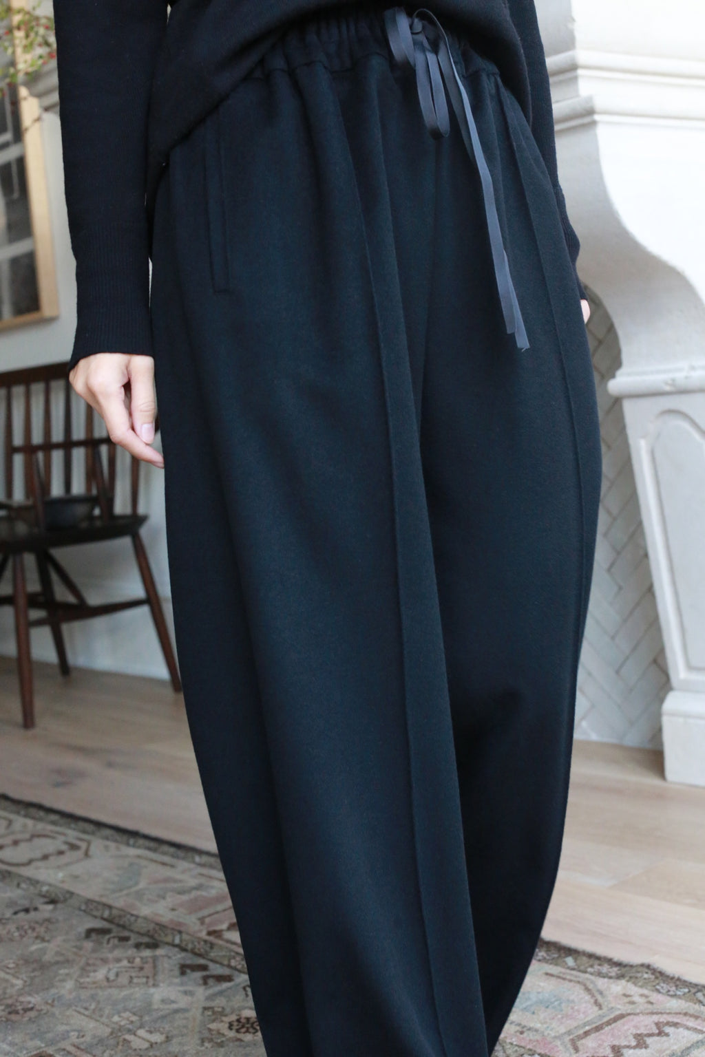 A person wearing loose-fitting, high-waisted, straight leg Cashmere Trousers with a drawstring waist stands indoors. The background features a cushioned chair, a wooden chair, and a portion of a fireplace, along with a patterned rug on the floor.