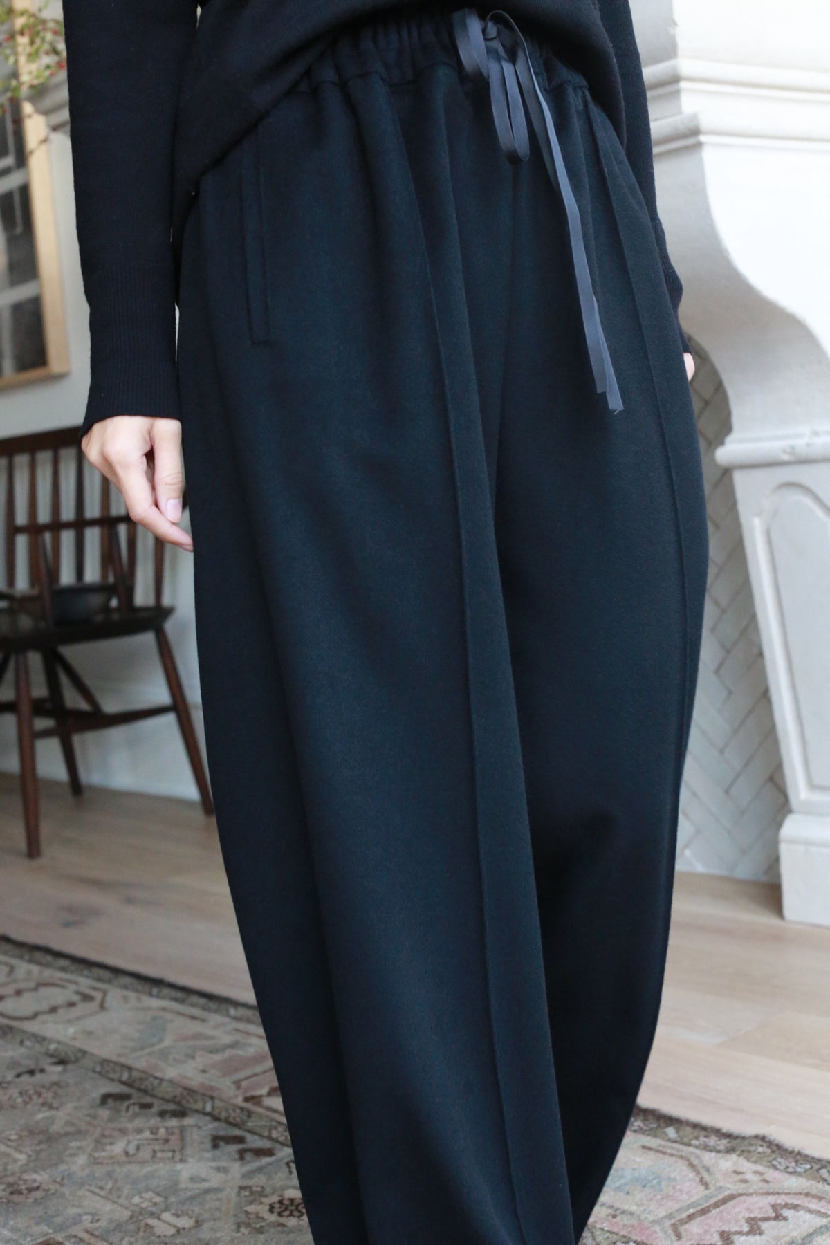 A person wearing loose-fitting, high-waisted, straight leg Cashmere Trousers with a drawstring waist stands indoors. The background features a cushioned chair, a wooden chair, and a portion of a fireplace, along with a patterned rug on the floor.