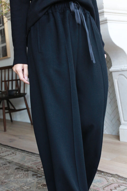 A person wearing loose-fitting, high-waisted, straight leg Cashmere Trousers with a drawstring waist stands indoors. The background features a cushioned chair, a wooden chair, and a portion of a fireplace, along with a patterned rug on the floor.