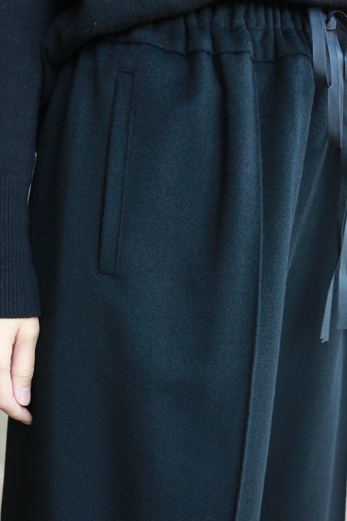 Close-up of a person wearing black Cashmere Trousers with an elastic waistband and a drawstring. Only the lower part of their sweater and their hand are visible. The pants have a side pocket.