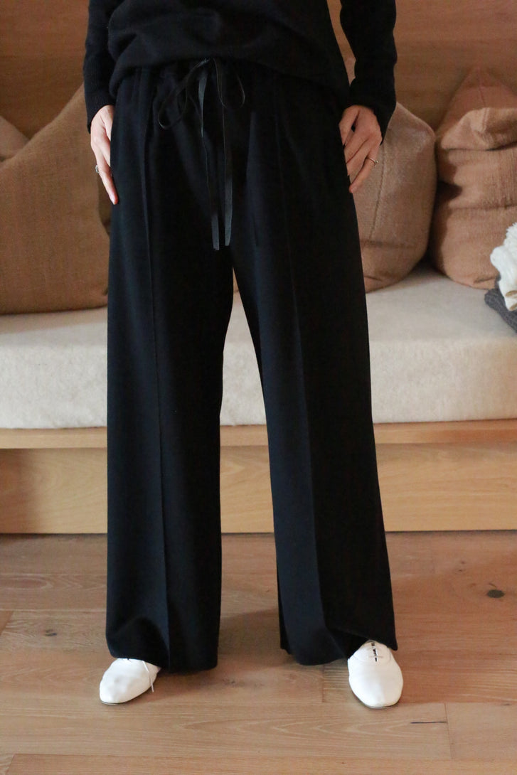 Person dressed in a black long-sleeved top and black Cashmere Trousers, standing on a wooden floor. White shoes are visible. A sofa with beige pillows is in the background.