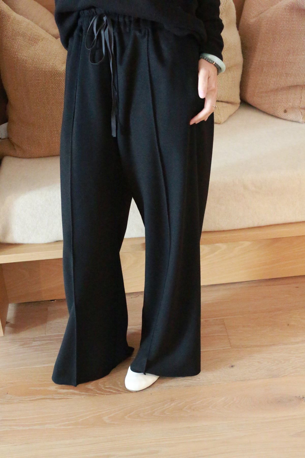 A person stands on a wooden floor wearing loose-fitting Cashmere Trousers with a drawstring waist and white shoes. The straight leg trousers add to their relaxed look. The person is positioned in front of a beige cushion on a light-colored, wooden bench, while their upper body is mostly out of frame.