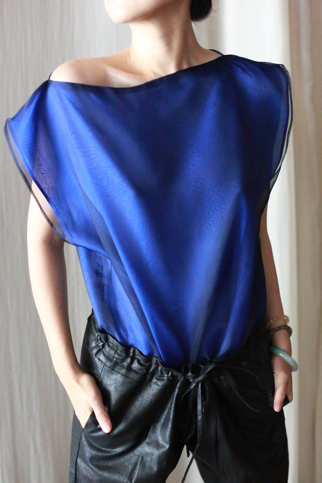 A person wearing a sheer blue top and Waxed Linen Unisex Pants with their hands in the pockets, standing against a light background. The head is slightly turned and the top exposes one shoulder.