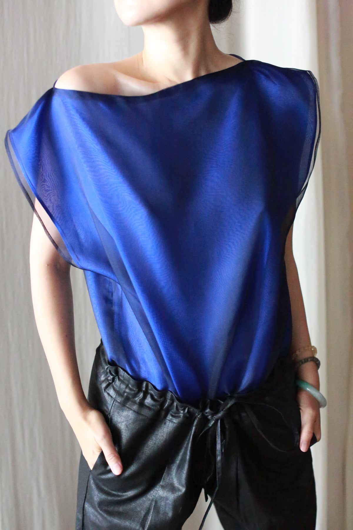 A person wearing a sheer blue top and Waxed Linen Unisex Pants with their hands in the pockets, standing against a light background. The head is slightly turned and the top exposes one shoulder.