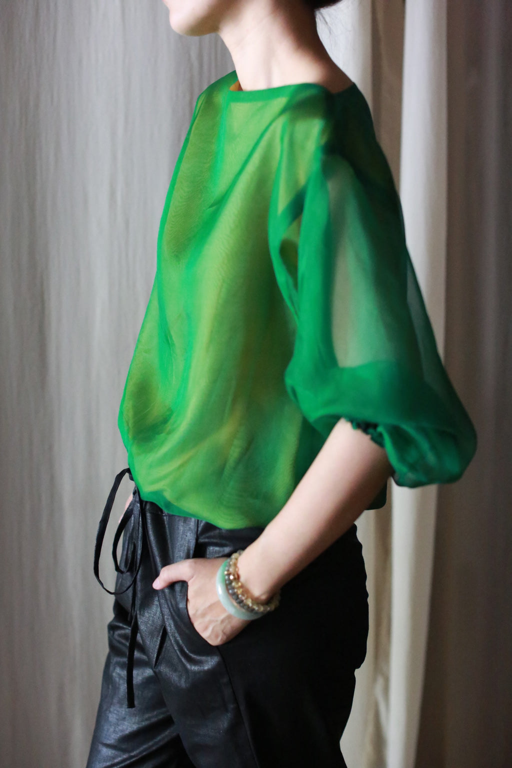 A person stands sideways with one hand in their pocket, dressed in a sheer green blouse with loose sleeves and black Waxed Linen Unisex Pants, accessorized with beaded bracelets.