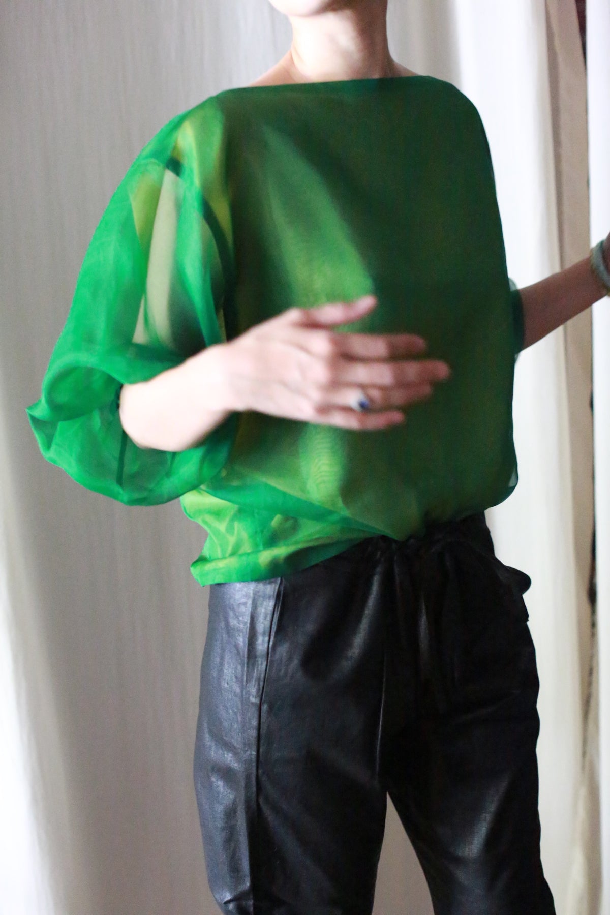 A person wearing a sheer green top with loose sleeves and Waxed Linen Unisex Pants stands against a light background. Their face is not visible, and one arm is slightly raised.