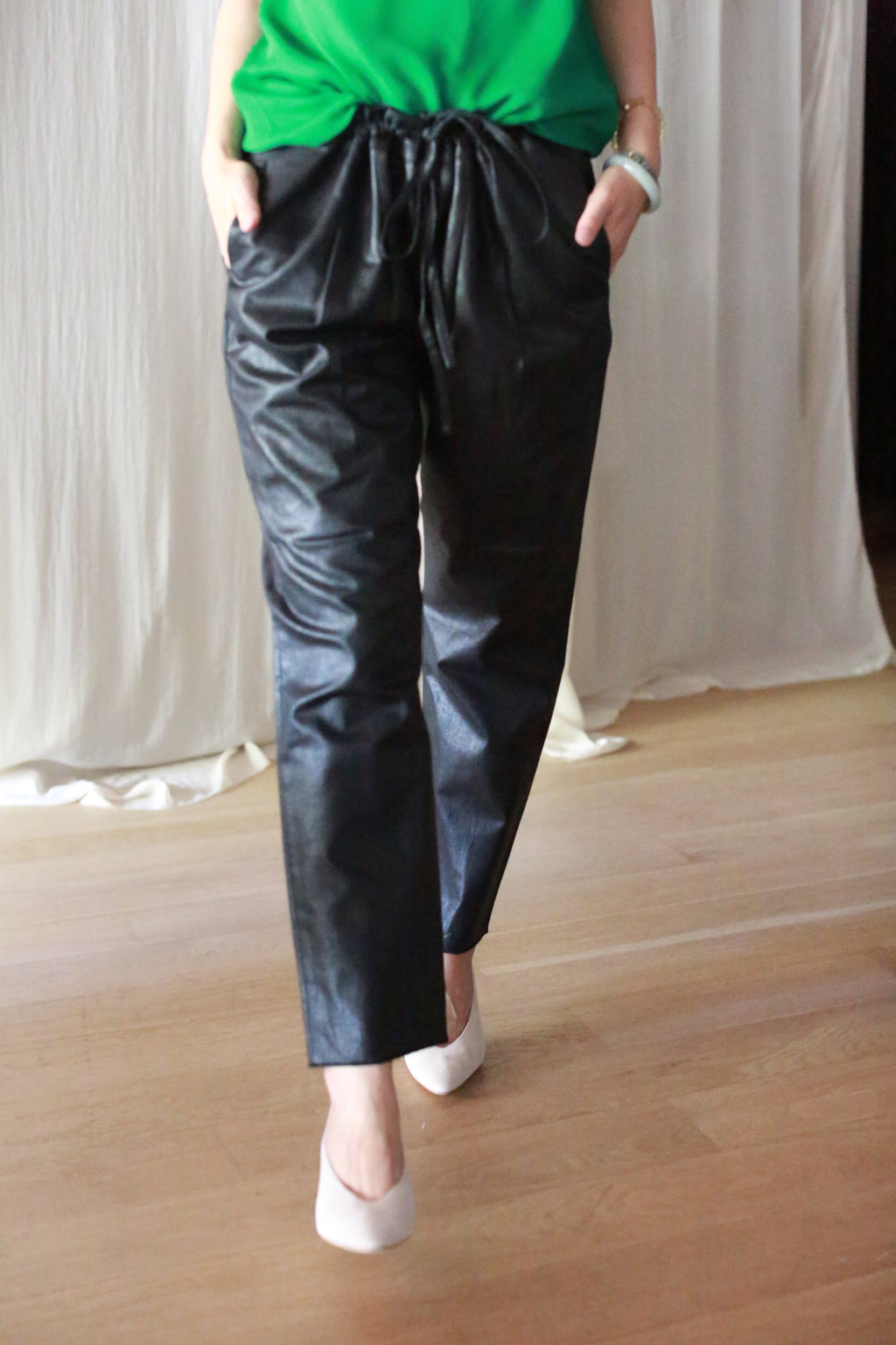 A person wearing black Waxed Linen Unisex Pants with a green top and white heels stands on a wooden floor in front of cream-colored curtains. Their hands are in the pants’ pockets, and their head is not visible.