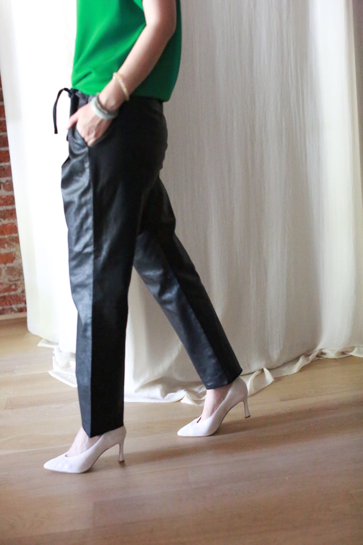A person in a green top, Waxed Linen Unisex Pants, and white heels walks indoors on a wooden floor with a white curtain and brick wall in the background.