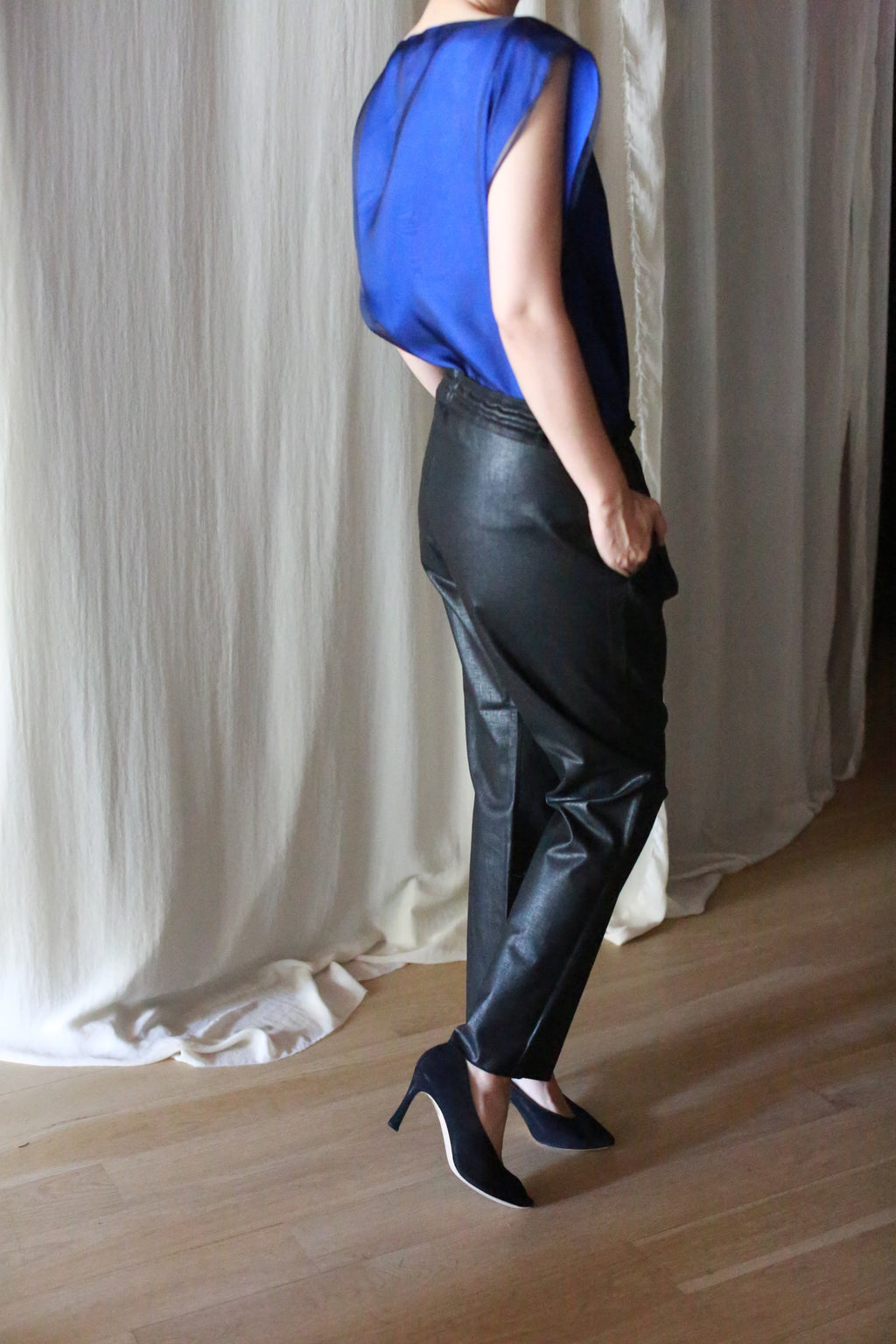 A person standing indoors, facing away, is wearing a sheer blue top, Waxed Linen Unisex Pants, and black pointed high heels. A light curtain backdrop and wooden floor are visible.