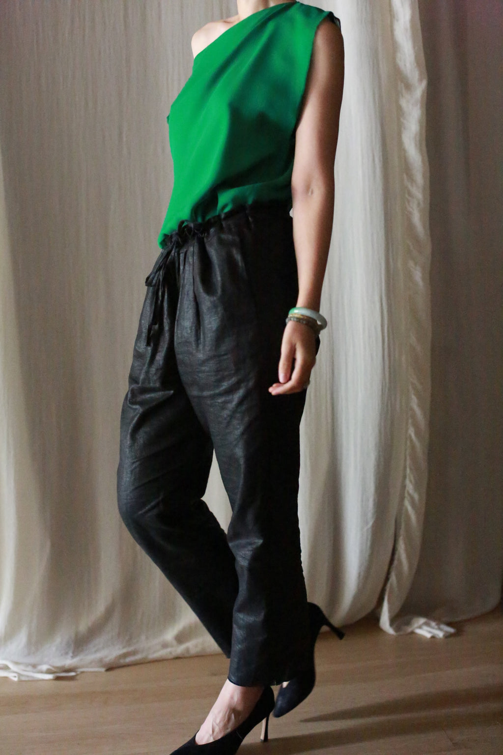 A person wearing a sleeveless green top and black Waxed Linen Unisex Pants with black heels, standing on light wood flooring in front of a cream-colored curtain.