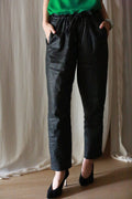 A person wearing Waxed Linen Unisex Pants in black, paired with a green top and black heels, stands with hands in pockets against a light, draped fabric background.