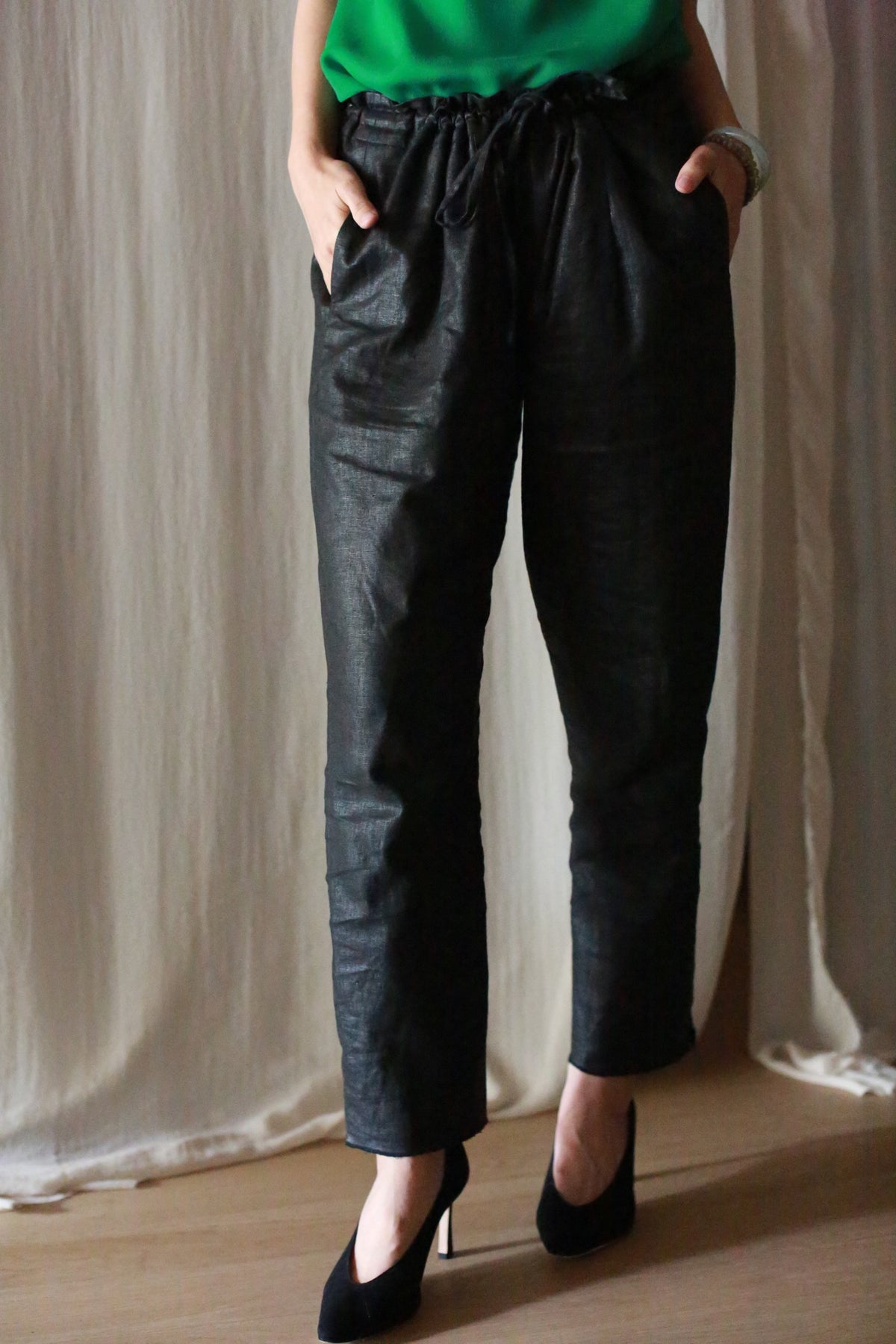A person wearing Waxed Linen Unisex Pants in black, paired with a green top and black heels, stands with hands in pockets against a light, draped fabric background.