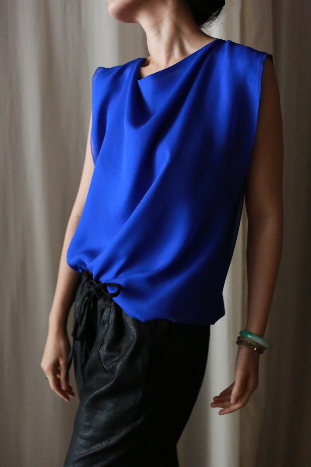 A person stands in front of a neutral background wearing Waxed Linen Unisex Pants and a sleeveless, bright blue blouse, with one arm slightly extended and head turned to the side.