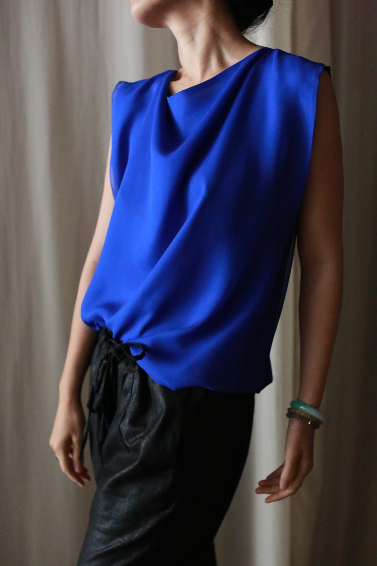 A person stands in front of a neutral background wearing Waxed Linen Unisex Pants and a sleeveless, bright blue blouse, with one arm slightly extended and head turned to the side.