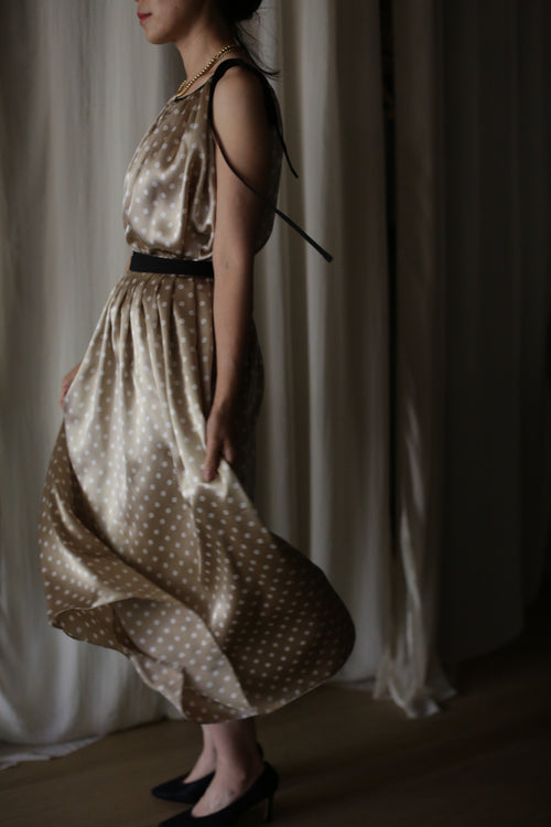A woman stands gracefully against a backdrop of draped white fabric. She is wearing a light brown, sleeveless dress with black straps and a black belt, adorned with ivory polkadots, paired with black heeled shoes. Her left hand holds part of the Charmeuse Pleated Wrap Skirt | Ivory Polkadot.