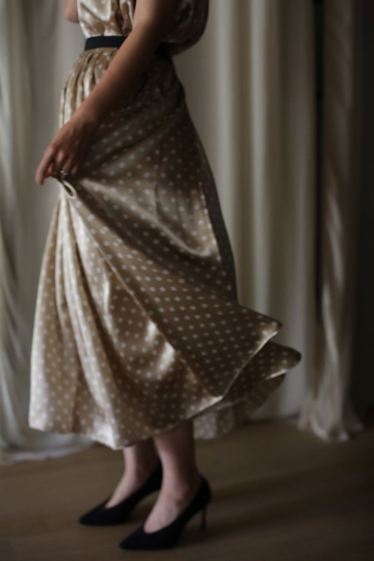 A woman stands indoors, holding the hem of her Charmeuse Pleated Wrap Skirt | Ivory Polkadot. She is wearing black high-heeled shoes. Light from a nearby window highlights the movement of the silk charmeuse fabric. The background features soft, blurred curtains.