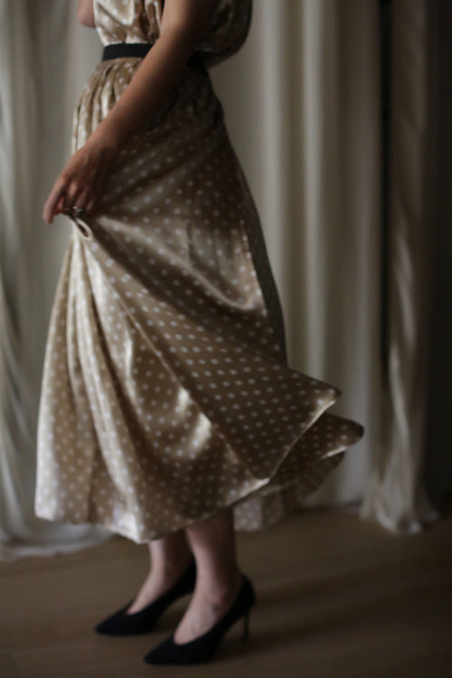 A woman stands indoors, holding the hem of her Charmeuse Pleated Wrap Skirt | Ivory Polkadot. She is wearing black high-heeled shoes. Light from a nearby window highlights the movement of the silk charmeuse fabric. The background features soft, blurred curtains.