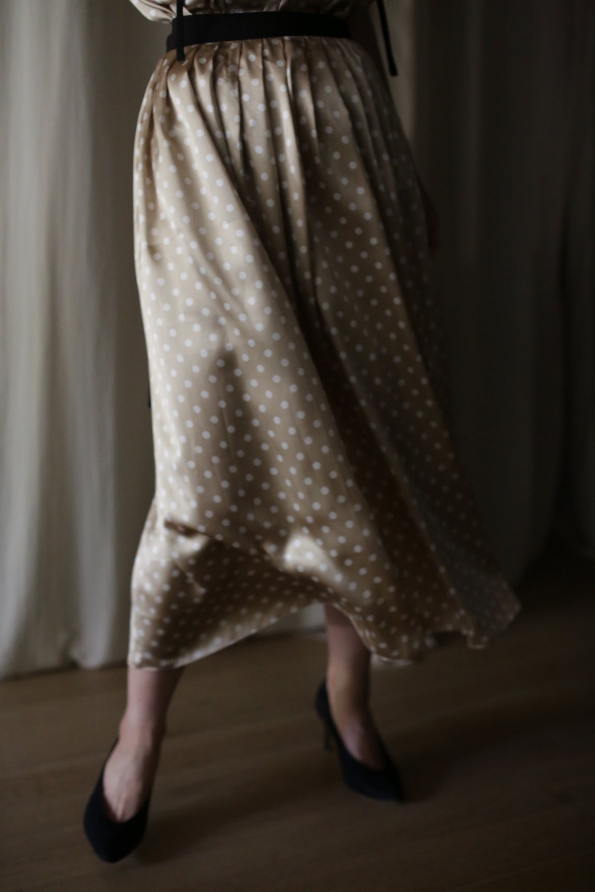 A person in a flowing, beige, silk Charmeuse Pleated Wrap Skirt | Ivory Polkadot dress is standing indoors. The photo captures the lower half of their body, showing off the skirt's elegant wrap design and their navy blue high heels. The background features light-colored, sheer curtains.