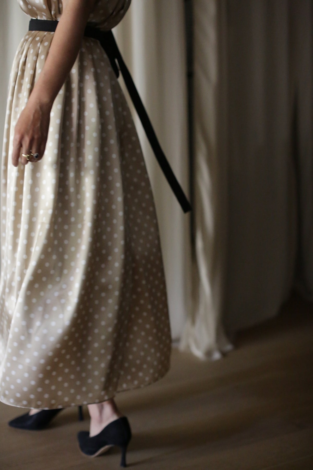 A person in a beige polka-dot dress with a black belt is standing indoors. The Charmeuse Pleated Wrap Skirt | Ivory Polkadot shimmers subtly, complementing their right hand adorned with rings and black high-heeled shoes. Only the lower half of their body is visible against a background of light-colored curtains and a wooden floor.