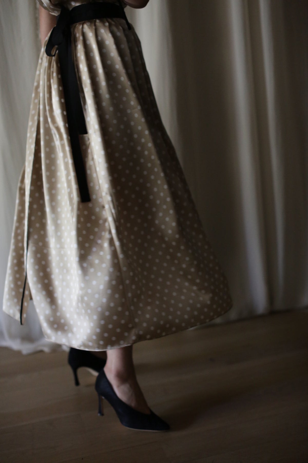A person wearing a Charmeuse Pleated Wrap Skirt | Ivory Polkadot with a black ribbon belt and black high heels stands on a wooden floor. The background features white curtains, adding a soft atmosphere. The image captures the lower half of the person's body.