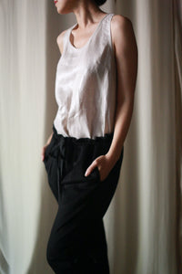 A person is standing with their hands in their pockets, wearing a sleeveless Stone Linen Tank and dark unisex pants. The background features a neutral, softly lit curtain. The individual’s head is turned slightly to the side, and their facial features are not visible.