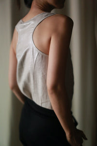 A person wearing the Stone Linen Tank and dark unisex pants is standing with their back to the camera. The soft lighting highlights the texture of the clothing and contours of their arm and shoulder. The background is blurred and neutral in color.