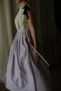 A person wearing a sleeveless white top with black ribbon details on the shoulders and a flowing, Typewriter Wrap Skirt | Amethyst with a tied waist ribbon. The image is taken indoors against a dark background, highlighting the elegant and classic outfit perfect for summer plans.