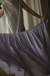 A person wearing a white top holds out the edges of a flowy light purple wrap skirt, creating a billowing effect. The image is softly lit, highlighting the Typewriter Wrap Skirt | Amethyst's delicate texture and movement. The background is neutral, keeping the focus on the clothing and evoking dreamy summer plans.