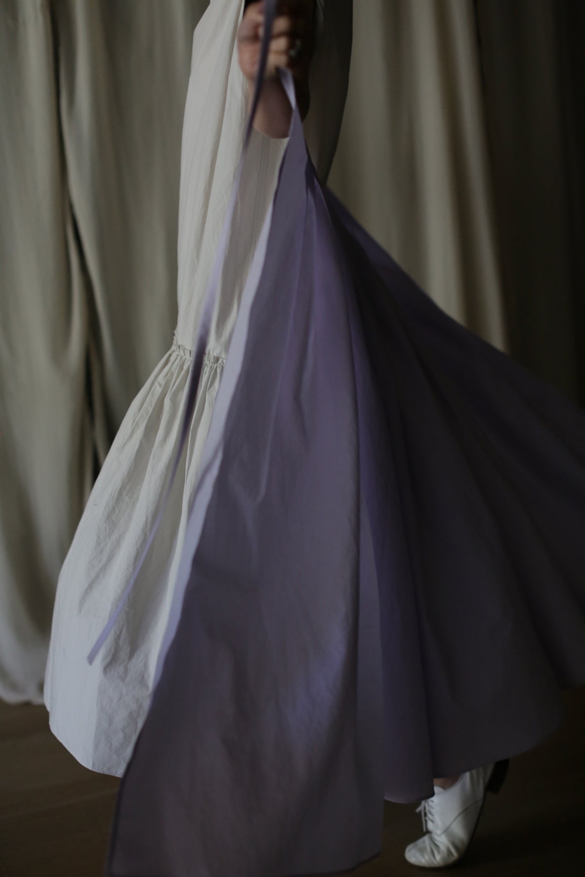 A person twirls while wearing a flowing Typewriter Wrap Skirt | Amethyst with white and lavender fabric. The image captures the motion of the skirt, with subtle lighting and an indistinct background. White shoes are also visible.