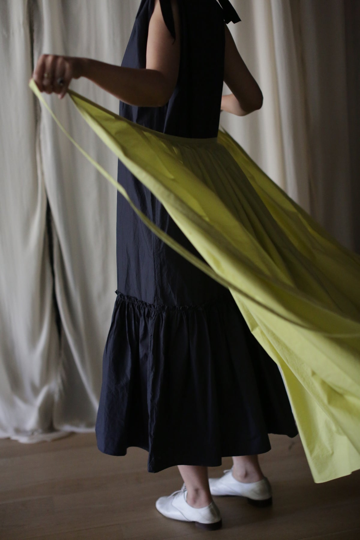 A person wearing a dark, sleeveless dress and white shoes is seen holding the ends of a large, flowing Typewriter Wrap Skirt | Citrine, creating a striking contrast against their outfit. The background features light-colored curtains.