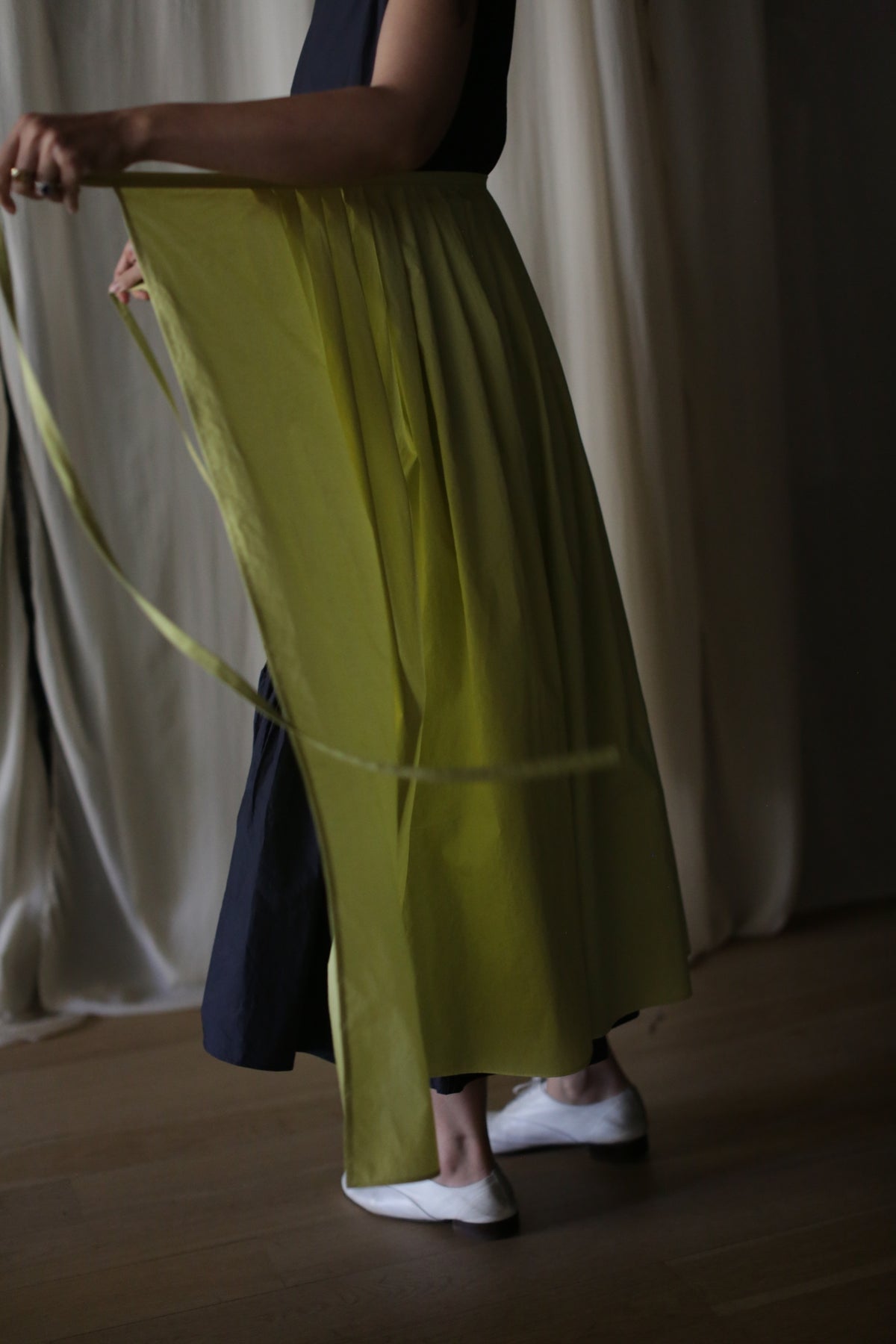 Person holding the edge of a Typewriter Wrap Skirt | Citrine with one hand, while the other hand holds a long ribbon. Standing indoors on a wooden floor with cream-colored curtains in the background, the lower half of the body is visible, wearing white shoes.