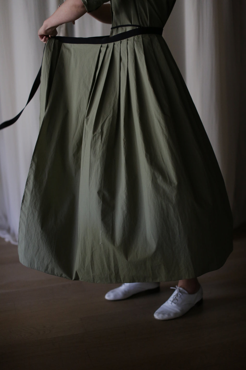 A person stands indoors on a wooden floor, their lower body visible in the Typewriter Wrap Skirt | Olive—an ankle-length, green pleated wrap skirt—paired with a black belt and white shoes.