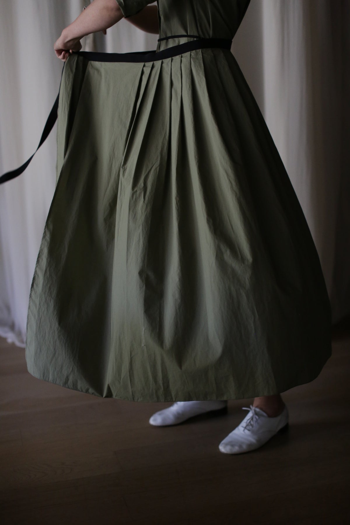 A person stands indoors on a wooden floor, their lower body visible in the Typewriter Wrap Skirt | Olive—an ankle-length, green pleated wrap skirt—paired with a black belt and white shoes.