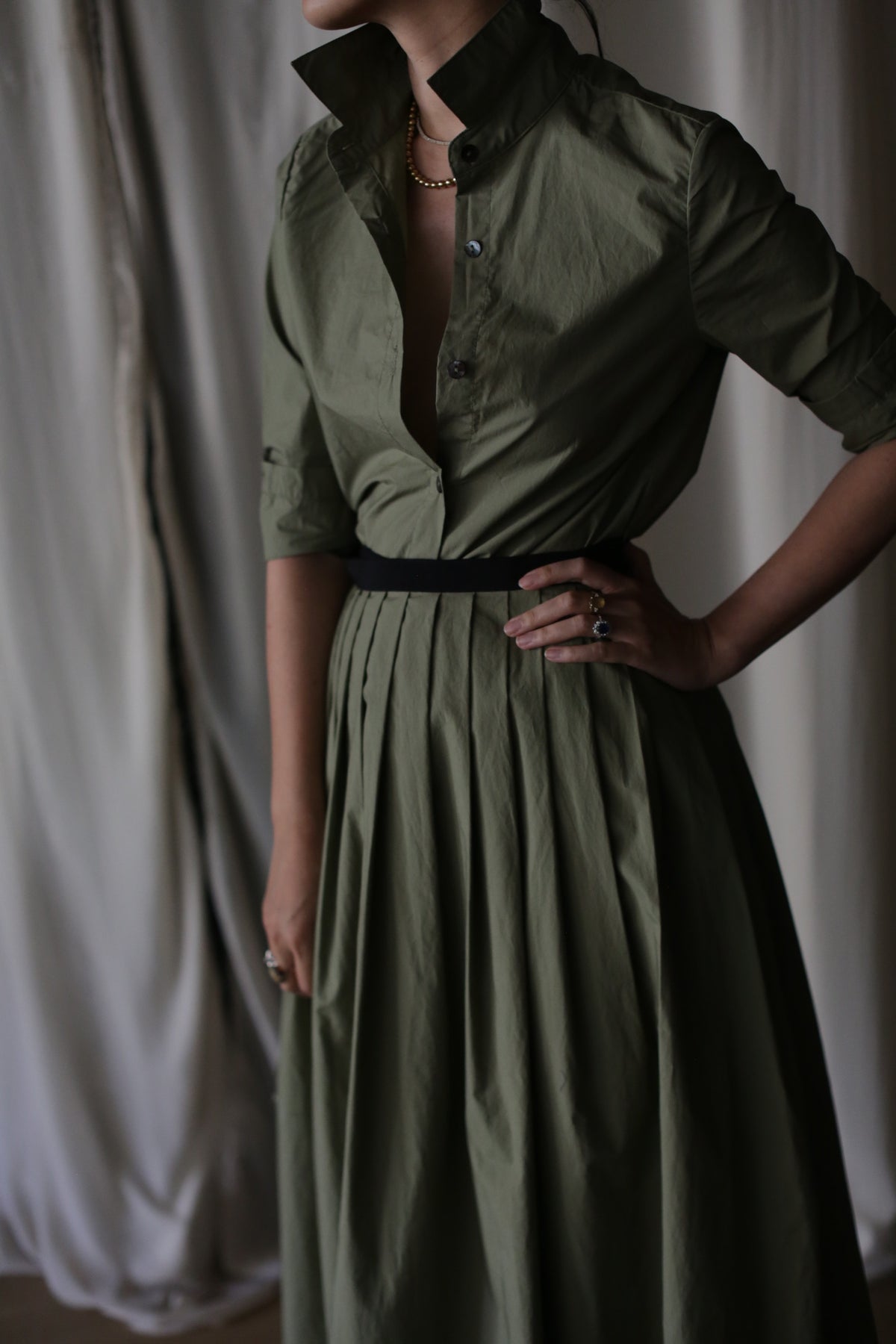 A woman stands with one hand on her hip, wearing the Olive Typewriter Wrap Skirt and a black belt. Her face is partially out of frame, and neutral curtains hang in the background.