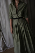 A person wearing the olive Typewriter Wrap Skirt with a black belt and pearl necklace stands before cream-colored curtains. The collared, buttoned long dress features a pleated skirt. Their face is not visible.